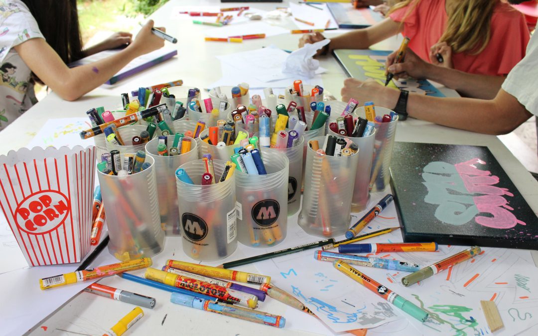 Tisch mit bunten Acrylstiften und zeichnenden Kindern